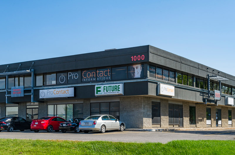 Le Carrefour du Commerce 1000 : immeuble commercial de deux étages, façade grise et noire, numéro 1000 en haut à droite, voitures stationnées devant.