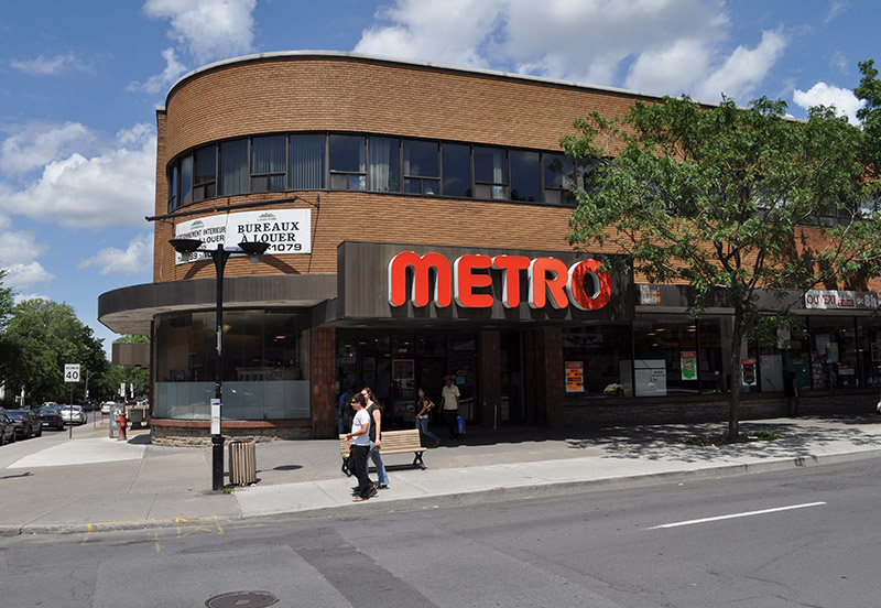 Le 5115, avenue Trans Island à Montréal : immeuble de brique arrondi à l’angle, épicerie au rez-de-chaussée, bureaux vitrés à l’étage.